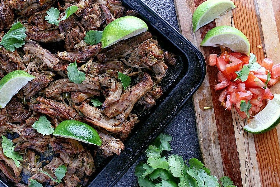 Freshly baked pork carnitas on a baking sheet with fresh limes and diced tomatoes