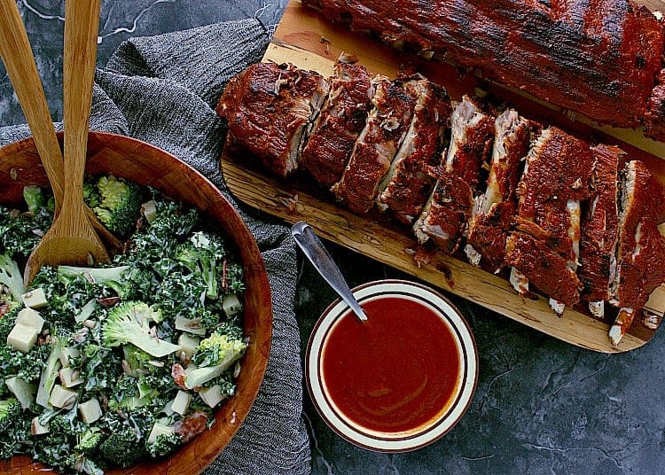 Two racks of ribs, one sliced into sections, next to a big bowl of broccoli salad and extra low carb barbecue sauce.