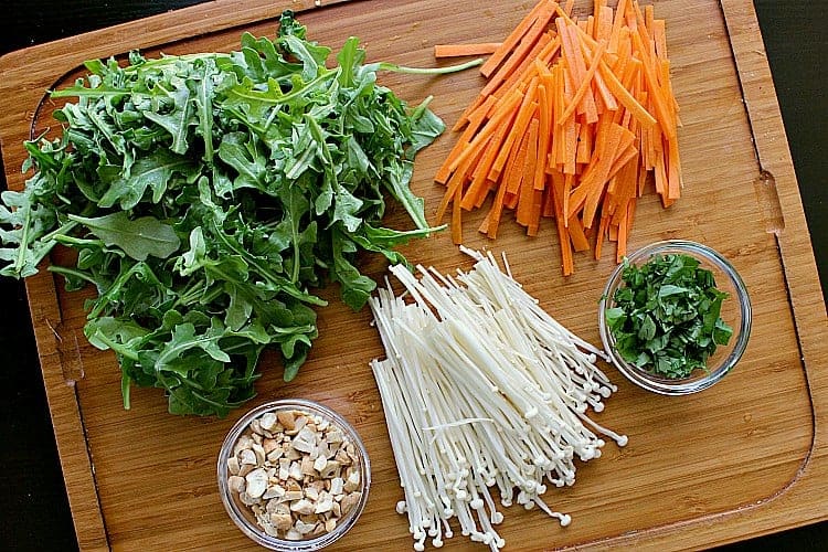 Cutting board filled with matchstick carrots, enoki mushrooms, cilantro, cashews and arugula.