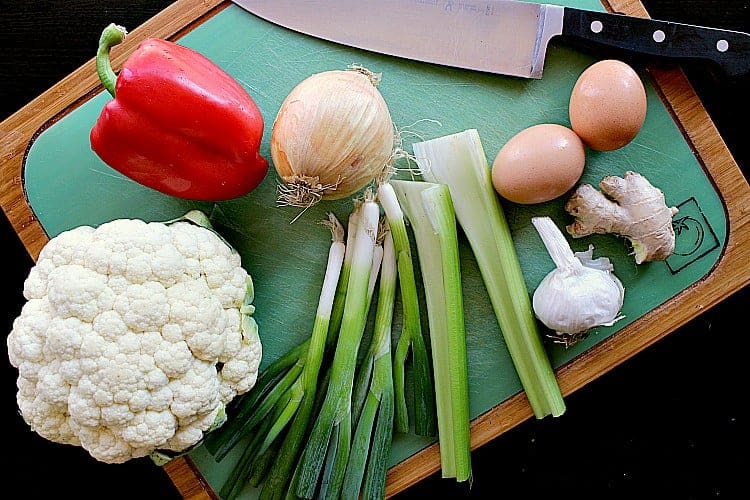 cutting board with cauliflower, red pepper, onion, green onion, celery, garlic, ginger and a couple of eggs.