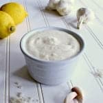 Bowl of garlic aioli next to some garlic, sea salt and lemons.