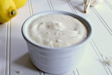 Bowl of garlic aioli next to some garlic, sea salt and lemons.