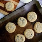 Baking sheet with 6 Keto chocolate chip cookies, next to a single cookie resting on a cutting board.