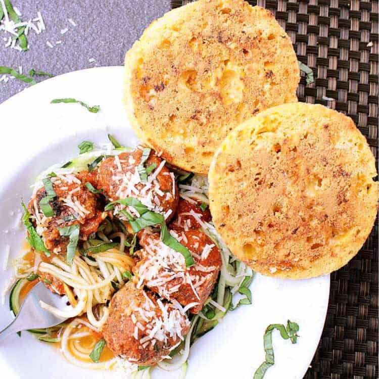 Two slices of keto garlic bread next to a bowl of low carb spaghetti with zoodles and meatballs. The dish is garnished with fresh parmesan and basil.