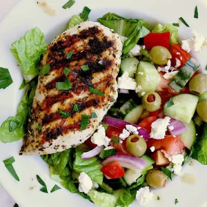 A white bowl with chopped romaine, greek salad and a piece of grilled keto greek chicken.