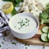 white bowl with keto ranch dressing, on a wooden cutting board with chopped vegetables.