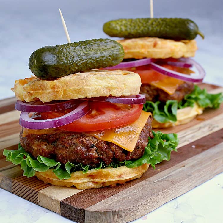 Two keto burgers loaded with cheese, tomato, red onion and lettuce, both on chaffles and placed on a wood cutting board.