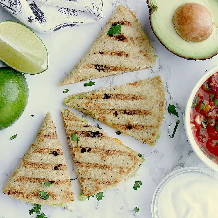 Keto Quesadilla sliced into 4 wedges next to lime wedges, half an avocado, a bowl of salsa and sour cream.