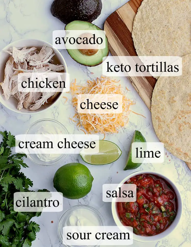 All ingredients used in this Keto Quesadilla - almond flour tortillas, an avocado, shredded chicken, shredded cheese, a lime, cream cheese, bunch of cilantro, salsa and sour cream.