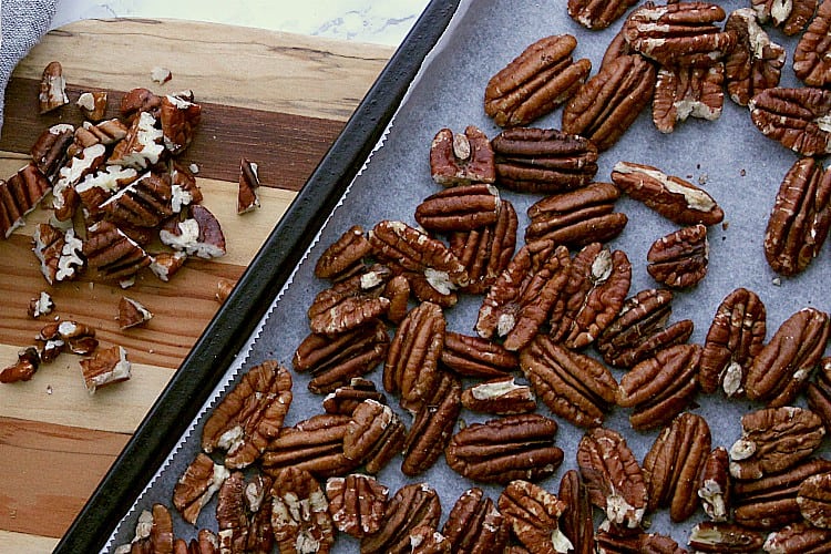 Roasted pecans on a baking sheet beside some chopped pecans.