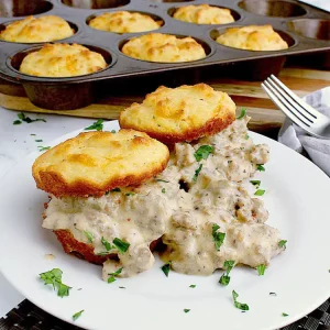 A plate with two biscuits, each cut in half and loaded with sausage gravy. The remaining tray of biscuits are behind the plate.