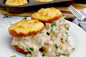 A plate with two biscuits, each cut in half and loaded with sausage gravy. The remaining tray of biscuits are behind the plate.