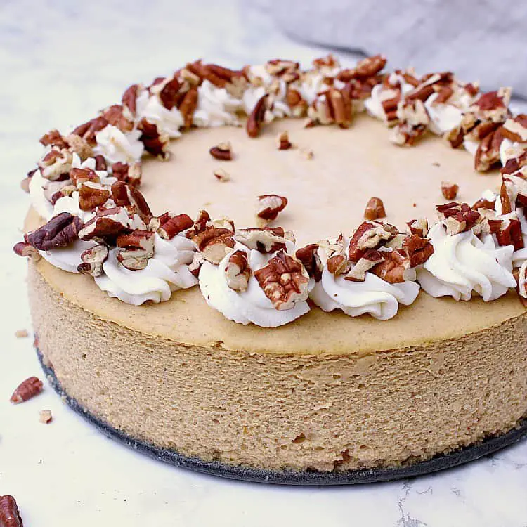 A Keto Pumpkin Cheesecake with the sides of the pan removed. The cheesecake is garnished with whipped cream and chopped pecans.