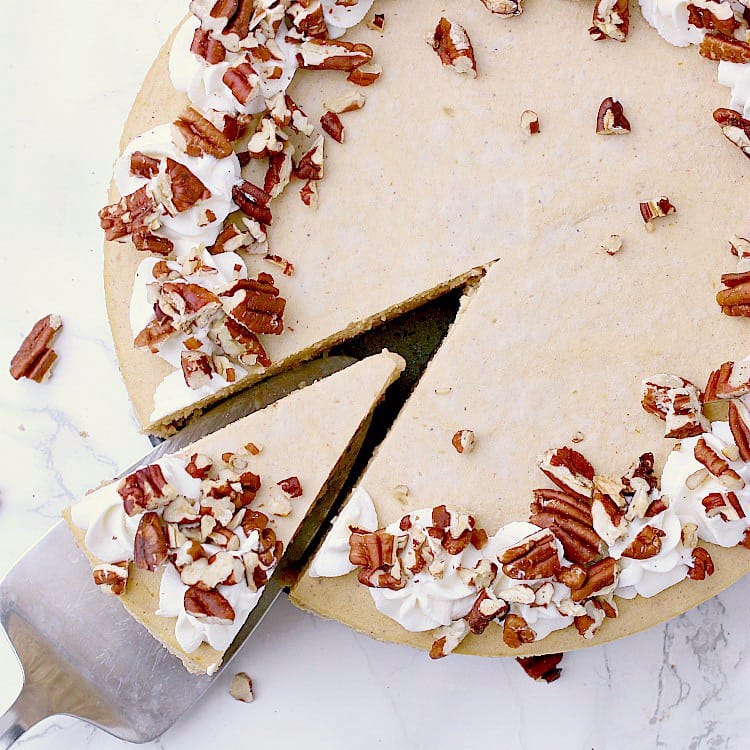 A Keto Pumpkin Cheesecake with the sides of the pan removed. The cheesecake is garnished with whipped cream and chopped pecans. One slice is being removed.