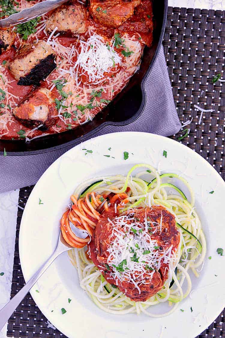 A bowl with zoodles, meatballs, parmesan and parsley, next to the cast iron dish with the cooked meatballs.