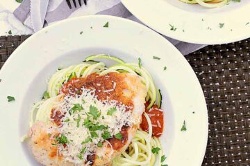 A white dish with zucchini noodles, marinara and chicken, topped with fresh parmesan and parsley.