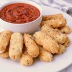 White plate with a stack of Keto Mozzarella Sticks and a bowl of Sugar Free Pizza sauce.