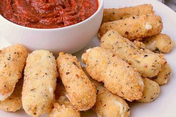 White plate with a stack of Keto Mozzarella Sticks and a bowl of Sugar Free Pizza sauce.