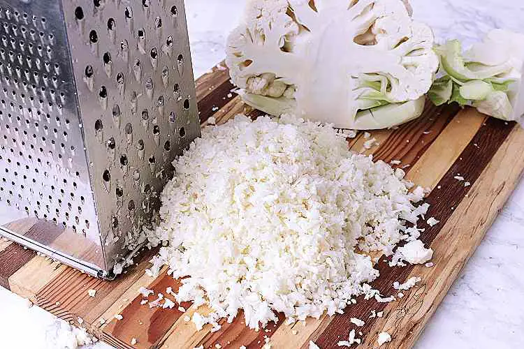 Cauliflower Rice on a wooden cutting board beside half a cauliflower and a cheese grater.