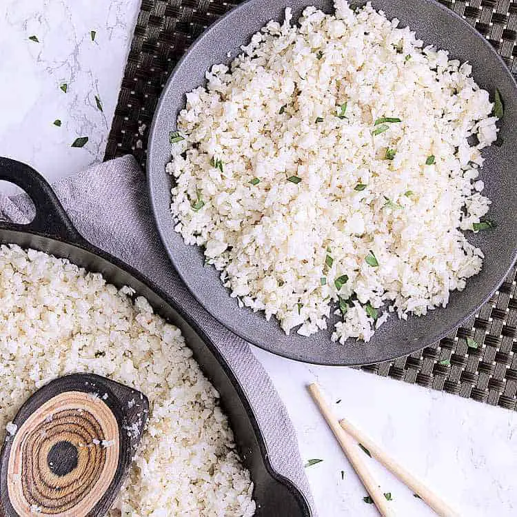 A bowl with cauliflower rice next to a cast iron skillet with cauliflower rice.