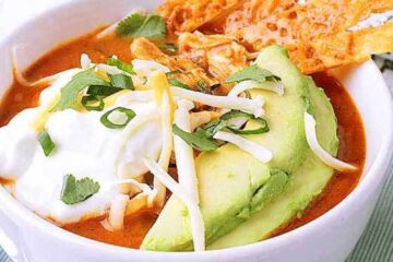 A white bowl with Keto Chicken Tortilla Soup topped with sour cream, cheese, avocado, cheese strips, cilantro and green onion.
