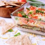 A serving dish with layered Keto Taco Dip beside a dish of chips.