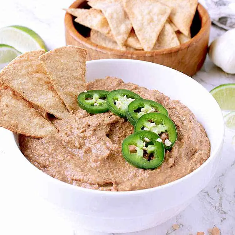 White bowl with low carb refried beans, garnished with sliced jalapeño and a few Keto chips. In the background there are sliced limes, garlic cloves and a bowl of keto chips.