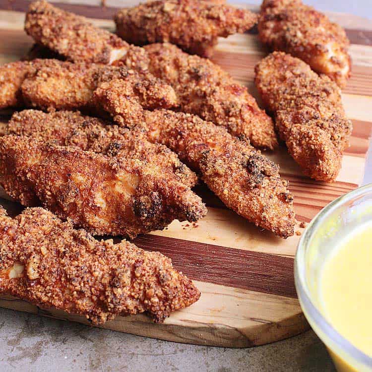 A wooden cutting board with crisp air fryer Keto chicken tenders, beside a small bowl of Keto honey mustard.